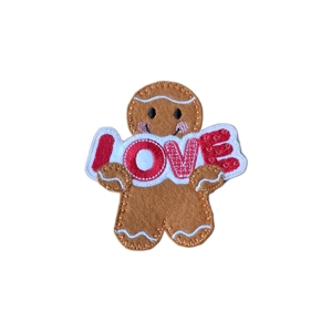 Gingerbread Love Character handmade from soft felt, holding a white stitched sign with the word “LOVE” in red letters. The gingerbread has pink cheeks, embroidered icing details, and a stitched smile.