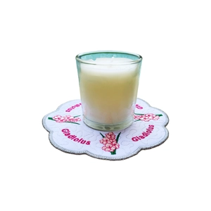 A white candle in a glass holder sitting on a scalloped embroidered mat featuring pink gladiolus flowers and the word “Gladiolus” stitched in matching pink around the design.