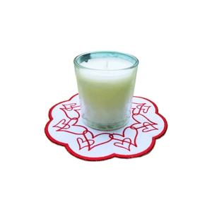 White candle in a clear glass holder placed on a round scalloped mat embroidered with looping red hearts on a white background with a red border.