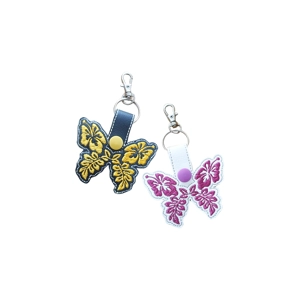 Two faux leather butterfly keyrings with hibiscus-style floral embroidery. One keyring is black with yellow stitching and a yellow snap, and the other is white with pink stitching and a purple snap. Both have silver keyrings and lobster clasps attached at the top.