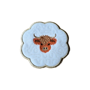 A scalloped candle coaster featuring an embroidered Highland cow with curved horns and shaggy fringe, stitched in warm brown and rust tones on a white fabric background, finished with a soft stitched border.