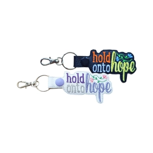 Two embroidered faux-leather keyrings with metal clasps, each reading “hold onto hope.” One is black with bright colourful stitching, and the other is white with pastel stitching and small floral details.