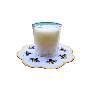 A white candle in a glass holder placed on a round embroidered table mat decorated with yellow and black honey bees.