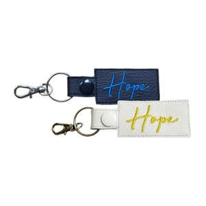 Two faux leather keyrings with the word 'Hope embroidered on them. One keyring is black with blue embroidery and a black snap fastener, attached to a silver keyring and silver lobster clasp. The other is white with yellow embroidery, a white snap fastener, and a silver keyring with a silver lobster clasp. Both keyrings are rectangular with textured stitching.