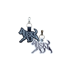 Two Husky silhouette keyrings with embroidered word-art spelling Husky, one in black with grey stitching and one in white with dark stitching, each attached to a metal keyring and clasp.