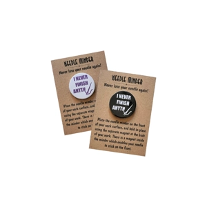 Two round 38 mm needle minders with the phrase “I Never Finish Anything” in bold lettering, displayed on brown kraft backing cards. One has purple text on a white background, and the other has white text on a dark background.