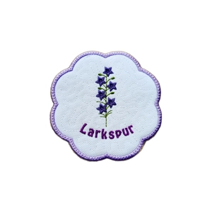 A scalloped embroidered candle coaster with a July larkspur flower stitched in purple and green, the word “Larkspur” embroidered below, on a white textured background with a stitched border.