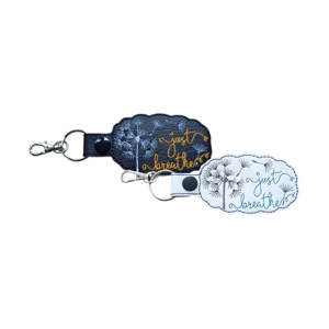 Two faux leather keyrings with embroidered dandelion designs and the words 'just breathe.' One keyring is black with gold stitching, and the other is white with blue stitching. Both have metal keyrings, lobster clasps, and snap fastenings.