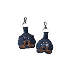 A kawaii-style Dachshund keyring made from black faux leather, featuring embroidered tan markings, floppy ears and cute sparkling eyes. Two versions are shown side by side: a short-haired Dachshund design on the left and a long-haired Dachshund design on the right, each with a metal keyring and lobster clasp.
