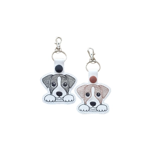 Two kawaii-style embroidered Jack Russell puppy keyrings shown side by side. One design is black and white with a black snap fastener, and the other is brown and white with a brown snap fastener. Both have cute puppy faces with big eyes, stitched paws and are attached to metal rings with lobster clasps.