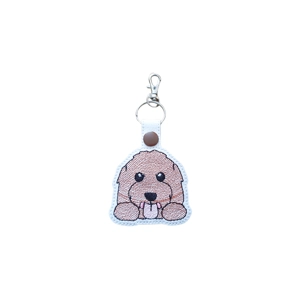 A white faux leather keyring featuring a cute embroidered Labradoodle with tan curly fur, big shiny eyes and a little tongue sticking out. The charm has a metal ring and clip at the top for attaching to bags or keys.