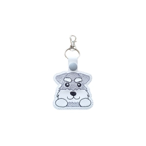 A faux leather keyring featuring a kawaii-style schnauzer pup with stitched details, including bright eyes, fluffy eyebrows and a little beard. The charm has a white snap tab, a metal keyring and a small lobster clasp at the top.