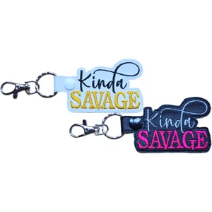 White and black faux leather Kinda Savage keyrings with colourful embroidered wording, split rings and lobster clasps, displayed side by side.