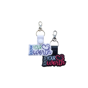 Two embroidered faux leather keyrings with metal clasps, each displaying the phrase 'Know Your Worth' in colourful stitched lettering with a heart. One keyring is white with blue, green and purple embroidery; the other is black with white and dark pink embroidery. They are shown hanging side by side against a plain background.