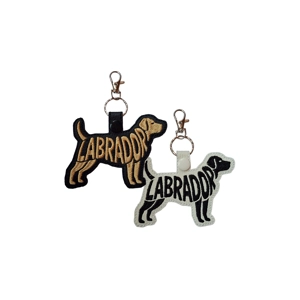 Two Labrador silhouette keyrings with embroidered word-art spelling Labrador, one in black with golden stitching and one in white with black stitching, each attached to a metal keyring and clasp.