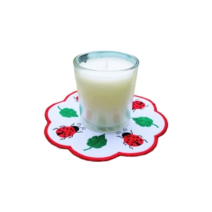 White candle in a clear glass holder placed on a round scalloped embroidered mat decorated with red ladybirds and green leaves on a white background with a red border.