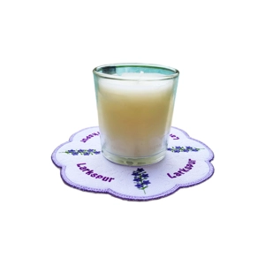 A white candle in a glass holder sits on a scalloped embroidered mat featuring purple larkspur flowers and the word “Larkspur” stitched in matching purple, representing the July birth flower.