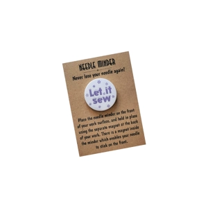 Round needle minder on a brown kraft backing card reading 'NEEDLE MINDER – Never lose your needle again!'. The minder is white with light purple text reading 'Let it sew', surrounded by small snowflake illustrations. The card includes instructions for using the magnetic needle minder for sewing or embroidery.