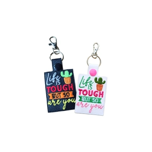 Two faux leather keyrings, one black and one white, machine embroidered with the words ‘Life Is Tough But So Are You’ and a cactus motif, finished with metal keyrings and lobster clasps.