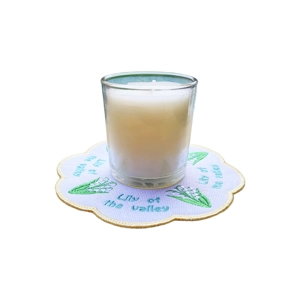 White candle in a clear glass holder placed on a round embroidered mat with lily of the valley flowers and text reading “Lily of the valley.”