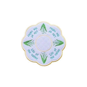 Round embroidered table mat with a scalloped edge, featuring green Lily of the Valley flowers and the words ‘Lily of the valley’ stitched in teal around a central stippling pattern.