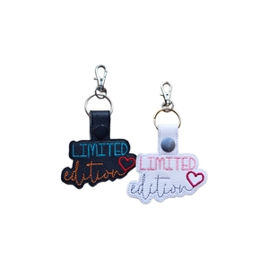 Two faux leather keyrings embroidered with the words 'Limited edition' and a small heart. One keyring is black with blue and orange stitching and a red heart, and the other is white with pink and grey stitching and a dark pink heart. Both have metal keyrings and lobster clasps.