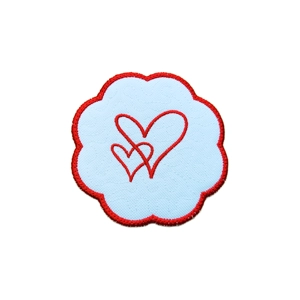 A white heart-shaped candle coaster with a red embroidered border and an interwoven heart motif stitched at the centre.