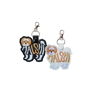 Two Malshi silhouette keyrings with embroidered word-art spelling Malshi and a cute stitched Malshi face, one in black faux leather and one in white, each attached to a metal keyring and clasp.