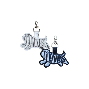 Two embroidered Maltese dog silhouette keyrings made from faux leather. One is white with grey stitching and a white snap fastener; the other is black with white stitching and a black snap fastener. Both feature the word 'Maltese' embroidered inside the dog outline and have metal swivel clasps for attaching to keys or bags.