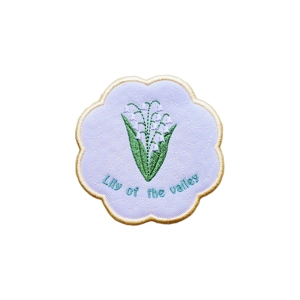 A scalloped embroidered candle coaster featuring lily of the valley stems stitched in green and white, with the words “Lily of the valley” embroidered below, on a white textured background with a stitched border.