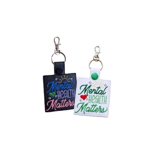Two faux leather keyrings embroidered with the words 'Mental Health Matters'. One is black with blue, green and pink stitching plus a yellow sun and small stars. The other is white with green and red stitching, a yellow sun and small stars. Both have metal keyrings and lobster clasps.