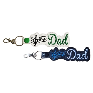 Two music-themed faux leather keyrings are shown. One is white with black musical notes and green embroidery. The other is black with blue musical notes and white embroidery. Both keyrings are attached to silver hardware.