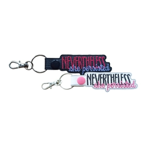 Two embroidered keyrings with metal clasps, each featuring the phrase “Nevertheless she persisted.” One keyring is made from black faux leather with dark pink and light purple stitching and a black snap fastener. The other is white faux leather with black and pink stitching and a pink snap fastener.