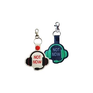 Black and white faux-leather keyrings featuring an embroidered gamer headset with the words Not Now. One is black with green stitching and grey text, and the other is white with black and red stitching. Each includes a snap tab, metal keyring, and lobster clasp.