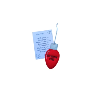 A red felt Christmas ornament shaped like a classic light bulb with “Christmas 2023” embroidered on the front in black thread. The bulb has a pale silver top and a light blue hanging loop. Behind it is a keepsake card titled “A Nurse’s Light,” printed with a heartfelt Christmas poem expressing appreciation for nurses.