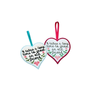 Two embroidered felt heart decorations featuring the quote “It takes a long time to grow an old friend.” One has a turquoise border with pink flowers and a teal ribbon, while the other has a deep pink border with pink flowers and a matching ribbon. Both hearts are white with stitched lettering and floral embroidery, representing friendship and lasting bonds.