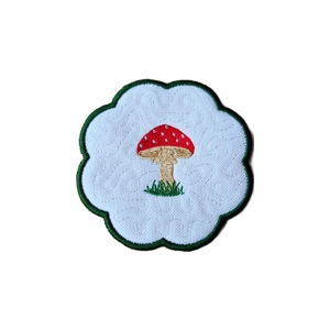 A scalloped fabric candle coaster featuring an embroidered red toadstool with white spots and a beige stem, set on a white stippled background with a dark green stitched border.