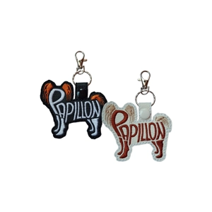 Two embroidered Papillon dog silhouette keyrings made from faux leather. One is black with white and orange stitching and a black snap fastener; the other is white with brown and cream stitching and a white snap fastener. Both feature the word 'Papillon' embroidered within the dog outline and include silver keyrings with swivel clasps.