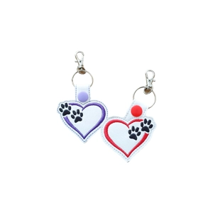 Two white faux leather keyrings shaped like hearts, each decorated with black pawprints. One has a purple heart outline with a matching purple snap, and the other has a red heart outline with a red snap. Both keyrings have silver rings and clasps attached.