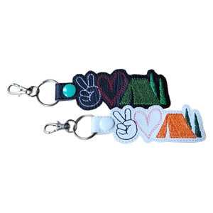 Two faux leather keyrings featuring Peace, Love & Camping designs. Each keyring shows a peace sign, a heart, and a pitched tent with trees. One keyring has a black base with white, red, and green stitching, and the other has a white base with black, pink, orange, and green stitching. Both include a metal key ring and lobster clasp.
