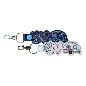 Two faux leather keyrings featuring Peace, Love & Caravan designs. Each keyring shows a peace sign, a heart, and a small caravan decorated with stitched details. One keyring has a black base with white, pink, blue, and grey stitching, and the other has a white base with black, pink, grey, and yellow stitching. Both include a metal key ring and lobster clasp.