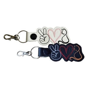 Two faux leather keyrings featuring a peace sign, a heart, and a sitting cat outline. One keyring has a black base with white, red, and orange stitching, and the other has a white base with black, pink, and black stitching. Each keyring includes a clasp and split ring. This design is called Peace Love Cat.