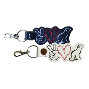 Two faux leather keyrings, one black and one white, each featuring stitched designs of a peace hand sign, a heart, and a dog silhouette. The black keyring has white and red embroidery with a black snap fastener, while the white keyring has black, red, and brown embroidery with a brown snap fastener. Both are attached to metal split rings and lobster clasps.