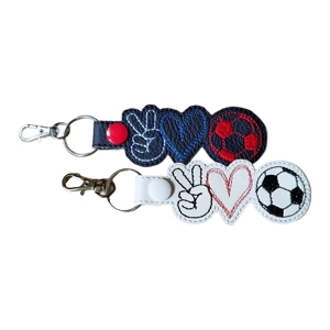 Two faux leather keyrings, one black and one white, each featuring stitched designs of a peace hand sign, a heart, and a football. The black keyring has white, blue, and red embroidery with a red snap fastener. The white keyring has black and red embroidery with a black-and-white football and a white snap fastener. Both are attached to metal split rings and lobster clasps.