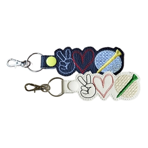 Two embroidered faux-leather keyrings are shown, each featuring three stitched symbols: a peace-sign hand, a heart, and a golf ball with a tee. One keyring is black with colourful stitching and a yellow snap; the other is white with contrasting threads and a white snap. Both have metal clasps and keyrings attached.
