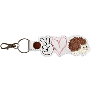 A white faux-leather keyring featuring embroidered designs of a black peace-sign hand, a pink heart, and a cute brown hedgehog. It has a brown snap button, a metal keyring, and a silver lobster clasp attached.