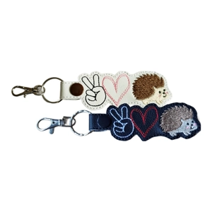 Two faux leather keyrings featuring a peace sign hand, stitched heart and hedgehog motif. One keyring is white and one is black, each finished with embroidered details and a metal ring and clasp.