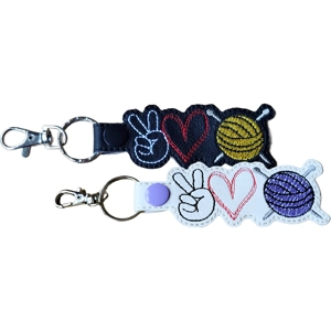 Two embroidered snap-tab keyrings, each shaped around three stitched icons: a peace-sign hand, a heart and a ball of yarn with knitting needles. One keyring is made from black faux leather with white, red and yellow stitching. The other is made from white faux leather with black, red and purple stitching. Both keyrings have silver hardware.
