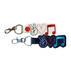 Two faux-leather keyrings with a Peace, Love, Music design. One is white with black, red, and red stitching, and the other is black with white, red, and blue stitching. Each features a peace hand sign, a heart, and a music note, along with a snap tab, metal keyring, and lobster clasp.