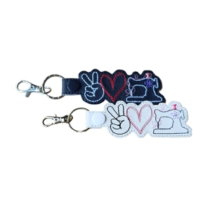 Two Peace Love Sewing keyrings shown together, one in black and one in white, each featuring an embroidered peace sign hand, stitched heart and sewing machine, finished with a metal ring and lobster clasp.